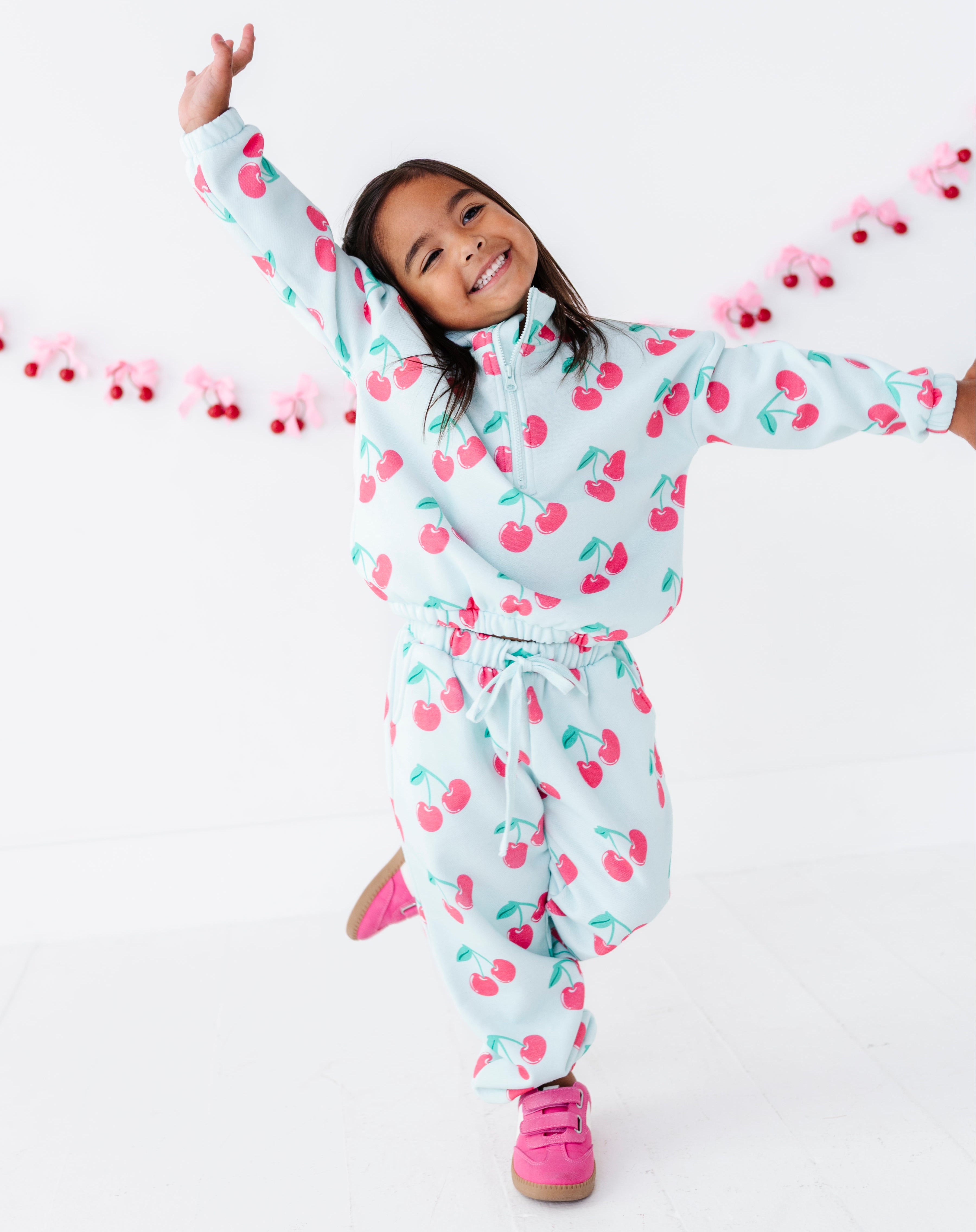 A young girl smiling and posing in cherry-patterned zip-up sweatshirt and matching sweatpants, with bright pink sneakers, against a clean white studio background with a pink cherry garland.